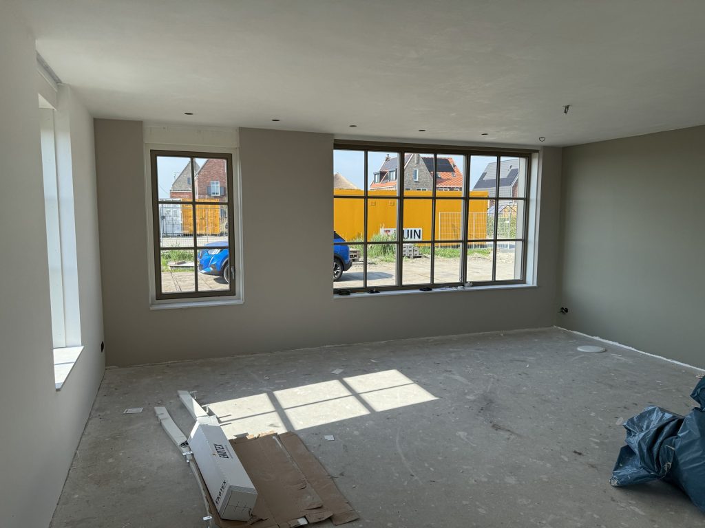 Woonkamer van een nieuw huis met grote roedeverdeelde ramen en warmgrijze wanden, nog zonder vloer