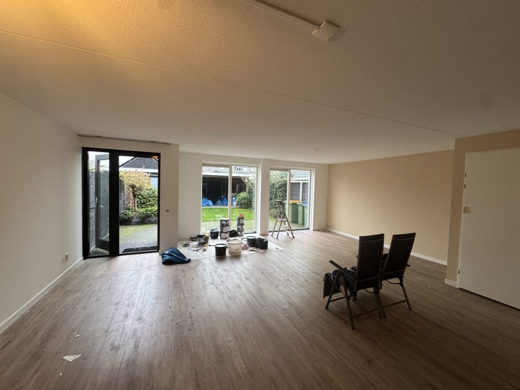 Ruime woonkamer van een bewoond huis met tuindeuren, laminaat en verfemmers bij het raam